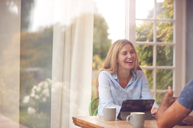 Echtpaar met digitale tablet zittend aan tafel werken vanuit huis bekeken door raam