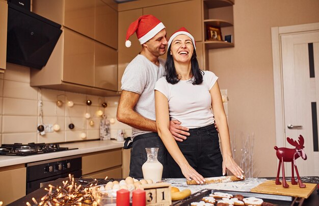 Echtpaar met kerstmutsen die samen binnenshuis op de keuken staan.