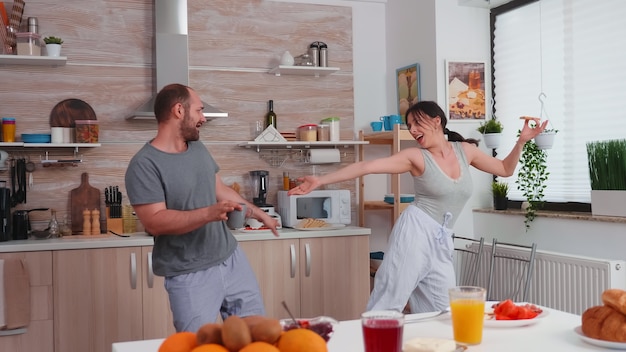 Echtpaar plezier dansen in de keuken tijdens het ontbijt. Zorgeloze man en vrouw lachen, zingen, dansen luisteren mijmerend, gelukkig en zorgeloos leven. Positieve mensen.