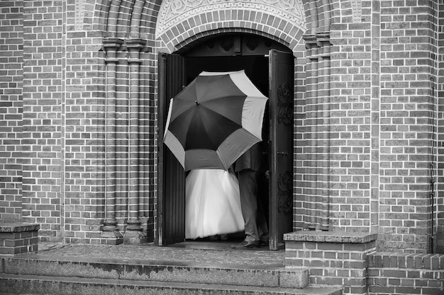 Echtpaar van bruid en bruidegom achter paraplu op kerkdeur op de dag van de huwelijksceremonie bij regenachtig weer