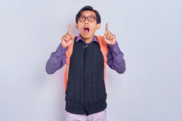 Ecited young asian student with a backpack with hands pointing
up to empty space isolated on gray background education in high
school university college concept