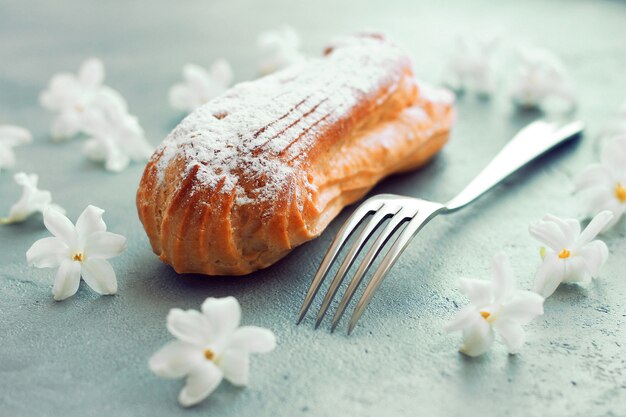 Eclair met poedersuiker op een grijze achtergrond, in de buurt van een vork, bloemen rond de compositie.