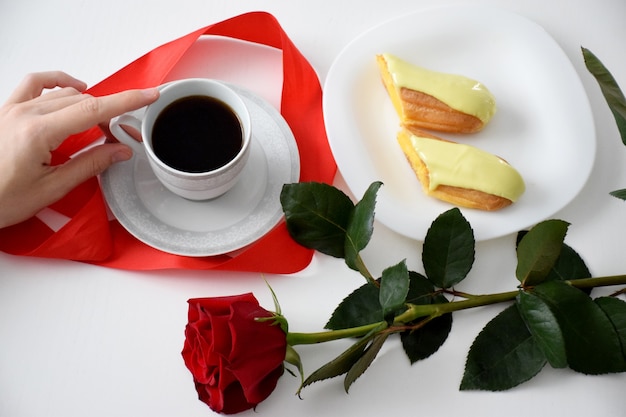 Eclair op een witte plaat, rode roos en kopje koffie. Valentijnsdag ontbijt.