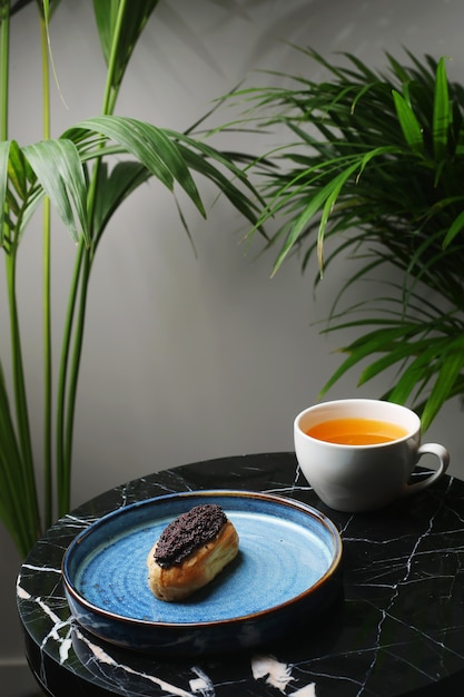 Eclair op keramische plaat en kopje thee op marmeren steen en tropische planten op achtergrond.