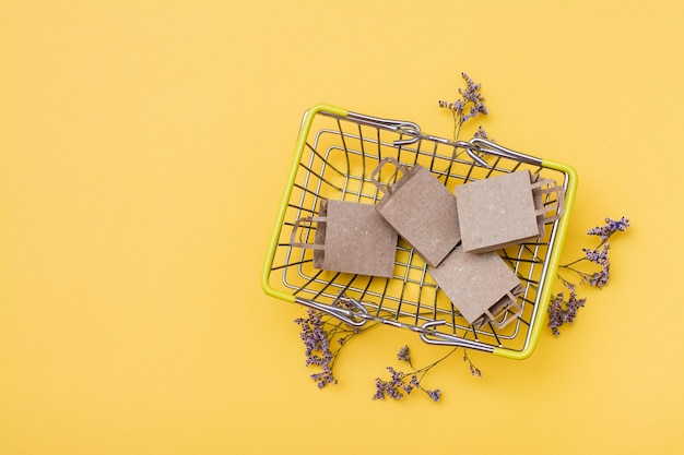 Eco-friendly craft paper bags in a metal shopping basket and dried flowers next to it on a yellow background. Black Friday, gift sales. Top view