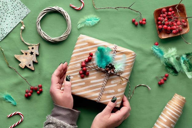 Photo eco friendly decorations - recycled craft wrapping paper, feathers, berry, wattle wreath, wooden christmas toys. hands wrapping xmas gift box with natural christmas decor on green textile tablecloth.