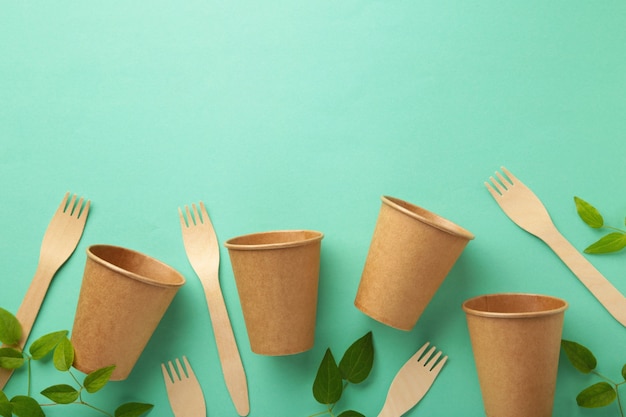 Eco friendly disposable glasses and fork with green leaves on mint background. Zero waste, eco friendly, plastic free background.