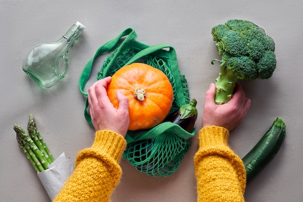 エコフレンドリーなゼロ廃棄物フラットブロッコリーとオレンジ色のカボチャのストリングバッグを両手で置いてください。野菜と手でフラット横たわっていた。