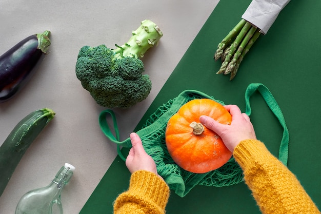 Piatto ecologico zero rifiuti a terra con mani in possesso di broccoli e sacchetto di stringa con zucca arancione. piatto disteso con verdure e mirtilli rossi in vaso di vetro su due colori di sfondo, carta marrone e verde.