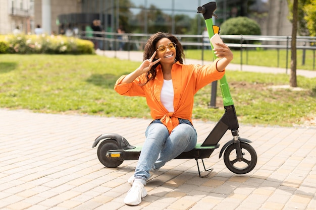 Eco personal transport in city glad millennial african american\
lady in casual taking selfie on smartphone