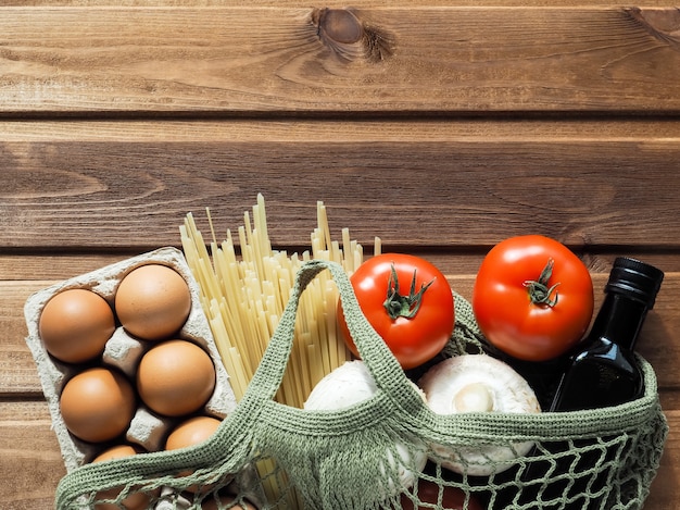 Eco-vriendelijke herbruikbare boodschappentas gevuld met eieren, pasta, tomaat, champignons en olie op houten achtergrond.