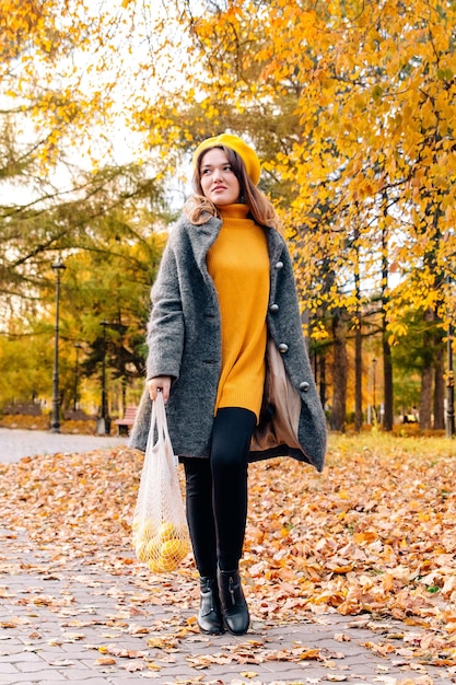 An ecobag with lemons is carried by a woman in an autumn park\
in a yellow sweater and coat