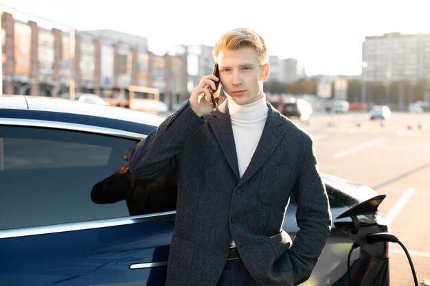 Ecological vehicles eco fuel concept close up portrait of\
handsome young blond man talking phone while his luxurious car is\
charging on city charging station