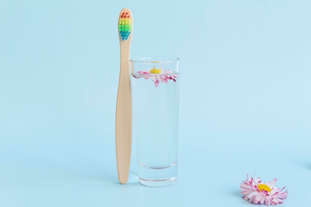 An ecological wooden toothbrush a glass with clean clear water and a white daisy floating in the water on a blue background