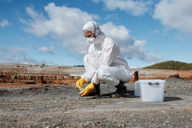 Ecologist in hazmat suit mask goggles and gloves gathering sample of soil on infertile land after en