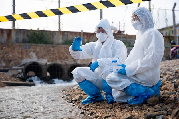 Эколог берет пробу воды из реки с помощью пробирки, ученый или биолог в защитном костюме и защитной маске собирает образец сточных вод из промышленных предприятий для анализа, проблемной среды