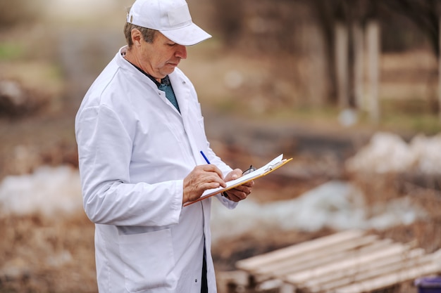 Ecologist in white uniform writing in clipboard results of land pollution while standing on landfill.