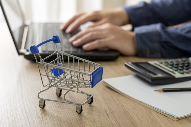 E-commerce e concetto di business online. carrello sulla tavola di legno e un uomo che utilizza un computer per chattare con il cliente in background.