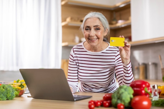 ECommerce Senior Woman With Laptop And Credit Card Posing In Kitchen Interior