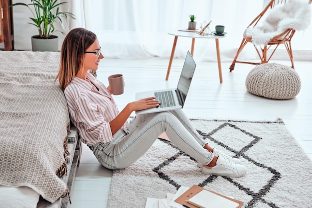 Женщина электронной коммерции Молодая леди, использующая компьютер для покупок в Интернете, сидит на полу и пьет кофе