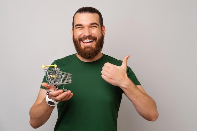 Ecstatic bearded man is holding a small trolley on his palm and his thumb up