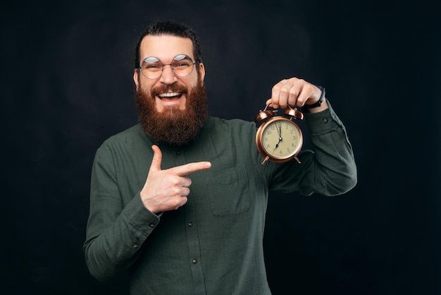 Ecstatic bearded man is pointing at round white wall clock while smiling Studio shot over black background