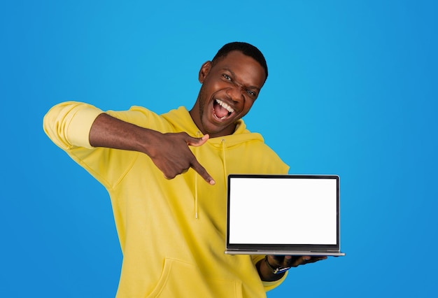 An ecstatic black man in a yellow hoodie points excitedly at a laptop screen with a blank display