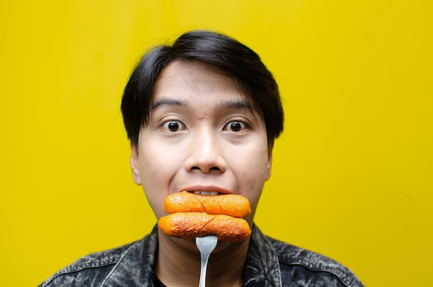 Ecstatic and expressive asian man eats and bites sausage using fork isolated over yellow background