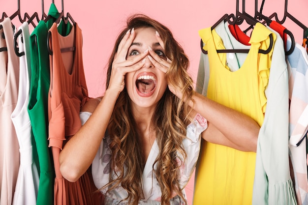 ecstatic woman in dress standing inside wardrobe rack full of clothes isolated on pink