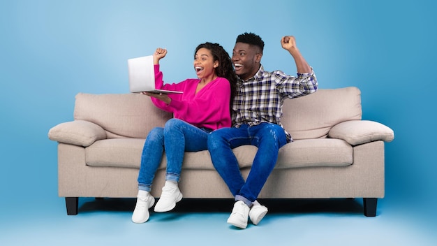 Ecstatic young black couple sitting on couch with laptop making\
yes gesture celebrating online win