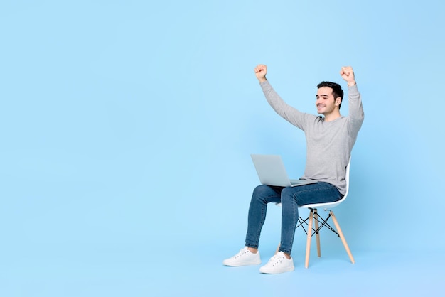 Ecstatic young winning Caucasian man raising both fists with laptop computer on the lap against light blue background with copy space
