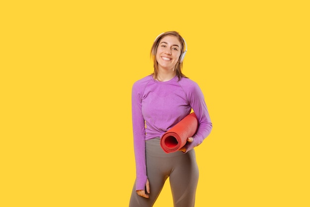 Ecstatic young woman is holding a red mat while wearing headphones