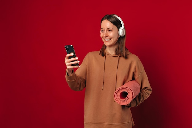 Ecstatic young woman wearing headphones is holding her fitness mat and phone