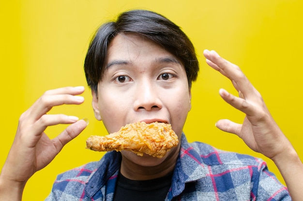 Ecstatische en expressieve Aziatische man eet en bijt gebakken kip geïsoleerd op gele achtergrond