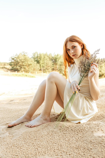 edhead vrouwelijk model in witte jurk zittend met grasboeket op zand