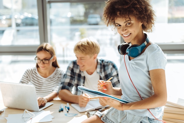 Educated generation. Positive smiling young afro american woman making notes and using headphones while her groupmates studying in the background