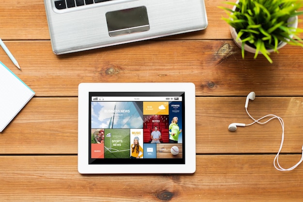 education, business, mass media and technology concept - close up of tablet pc computer, laptop and earphones on wooden table with internet application on screen