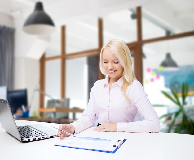 education, business and technology concept - smiling businesswoman or student with laptop computer and papers sitting over office room background