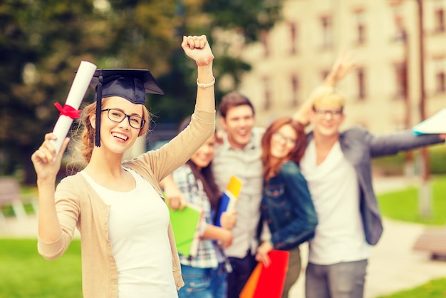 education, campus and teenage concept - smiling teenage girl in corner-cap and eyeglasses with diploma and classmates on the back