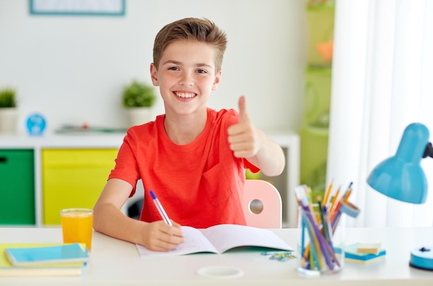 Education childhood and people concept happy student boy writing to notebook at home and showing thumbs up