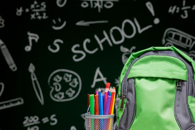 Education concept - green backpack, notebooks and school supplies by a blackboard