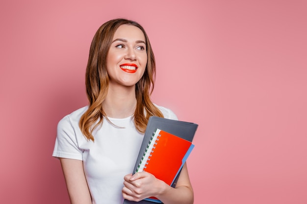 Concetto di educazione. felice studentessa caucasica che sorride distoglie lo sguardo e tiene quaderni, cartelle di libri in mano isolate su sfondo rosa in studio