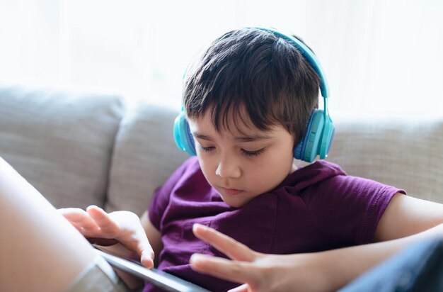 Education conceptHigh key portrait primary school Kid reading Ebook on tablet for homeworkChild wearing headphone playing game online on internetboy sitting on sofa watching cartoon on digital pad