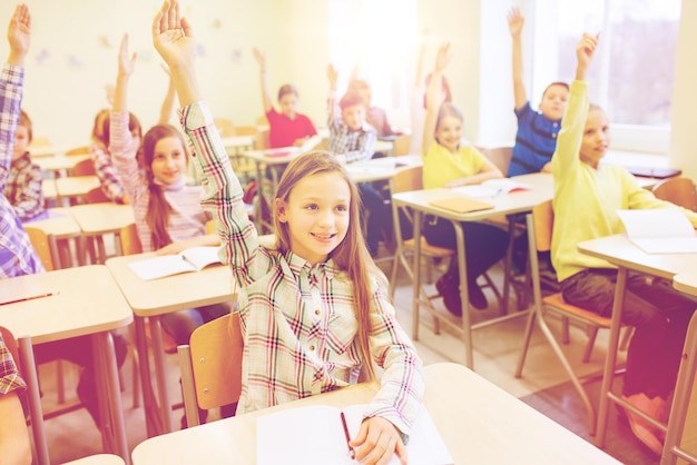 写真 教育、小学校、学習、人々の概念-教室に座って手を上げるノートを持った学校の子供たちのグループ