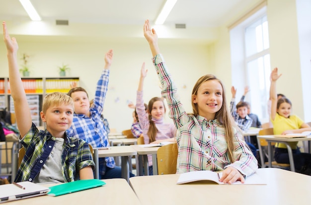 教育、小学校、学習、人々の概念-教室に座って手を上げるノートを持った学校の子供たちのグループ