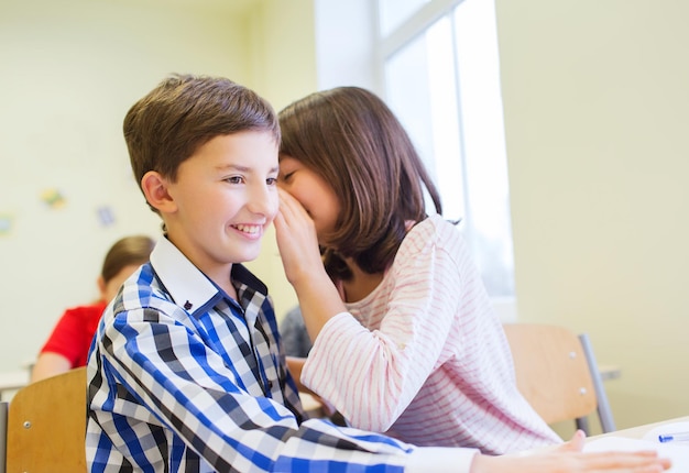 Concetto di istruzione, scuola elementare, apprendimento e persone - studentessa sorridente che sussurra il segreto all'orecchio del compagno di classe in classe