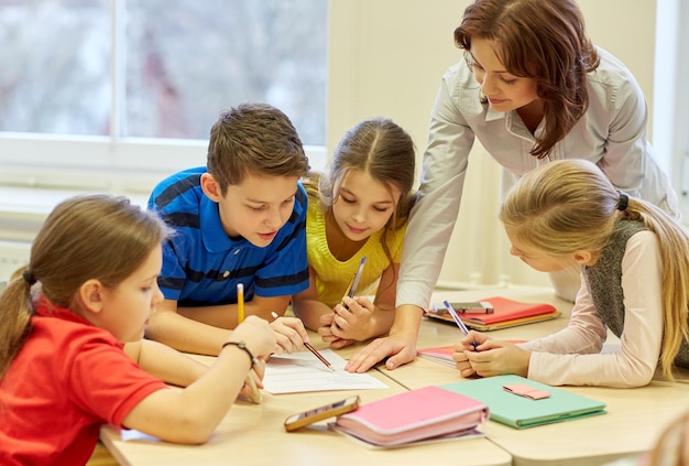 education, elementary school, learning and people concept - teacher helping school kids writing test in classroom