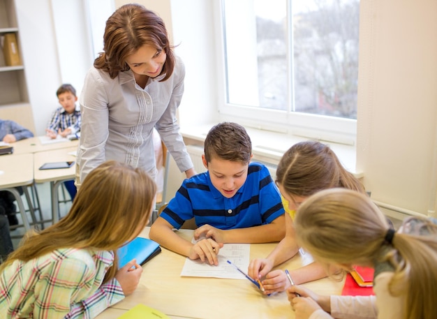 education, elementary school, learning and people concept - teacher helping school kids writing test in classroom