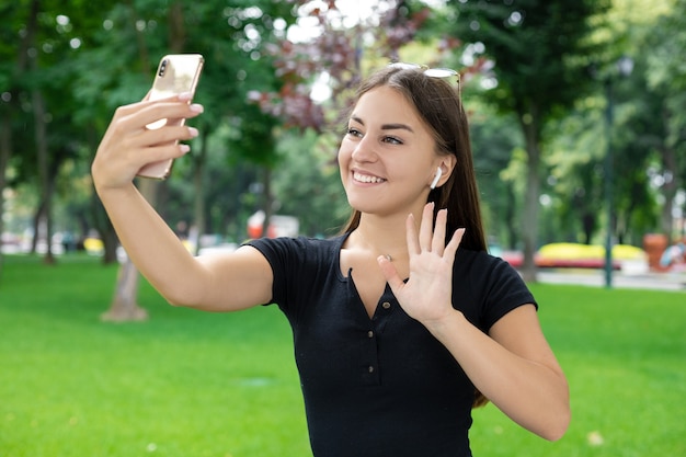 Education, freelance, fresh air, business, technology, and Internet concept â Smiling European-looking girl wearing eyeglasses and wireless headphones waves âhelloâ by video link into her phone.