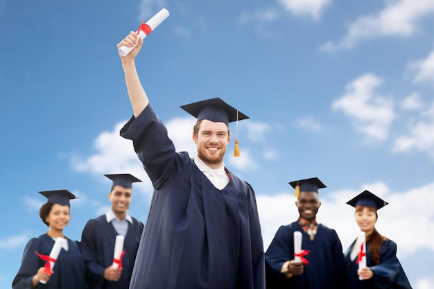 education, graduation and people concept - group of happy international students in mortar boards and bachelor gowns with diplomas over blue sky and clouds background